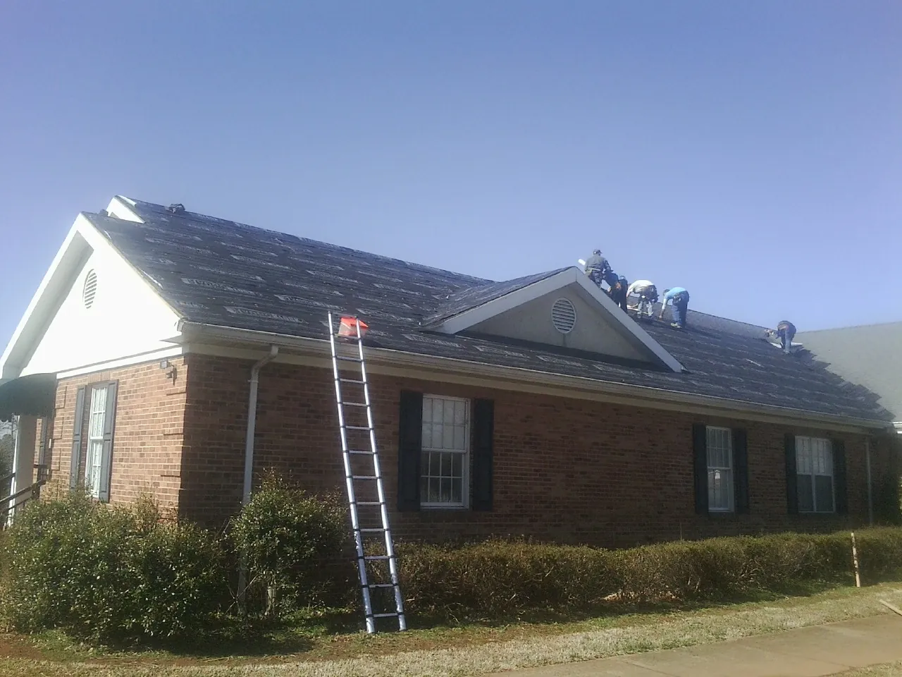 Crew tearing shingles off a brick ranch roof with a ladder set — Metal Roof Repair work by our crew in Elwood