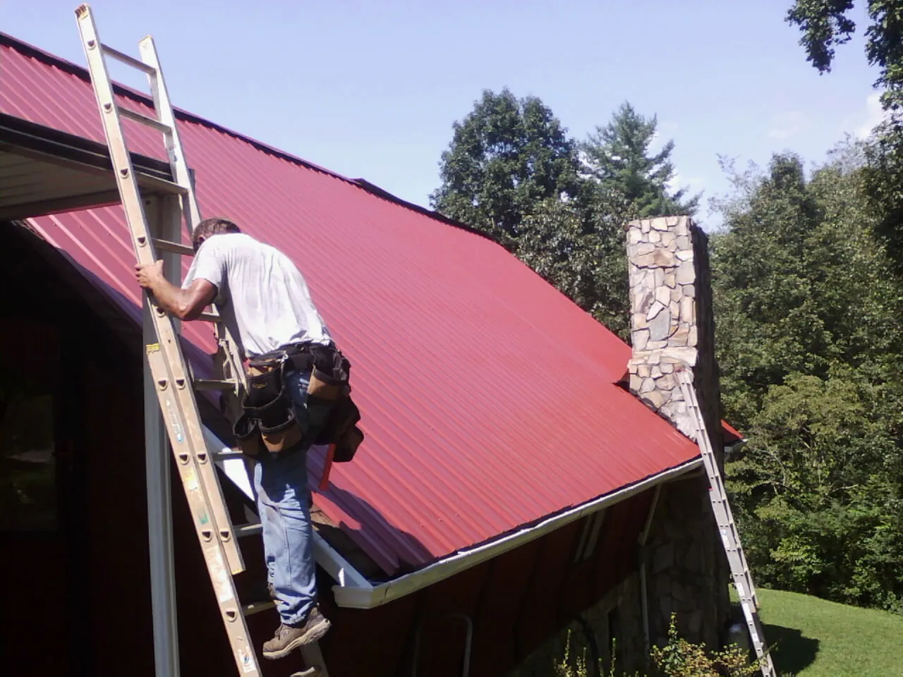 Roofer on a ladder beside a red metal roof and stone chimney — Soffit & Fascia Repair by Roofing Brothers in Elwood